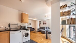 A modern kitchen with a dishwasher, washing machine, and microwave. There is a dining area with a table and chairs, and a sofa. The room has wooden flooring and a large window.