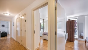 A hallway with wooden flooring, white walls, and a view into a bedroom with a bed and a window.