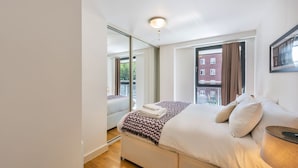 A modern bedroom with a large bed, a bedside table, a mirror, and a view of a red brick building through the window.