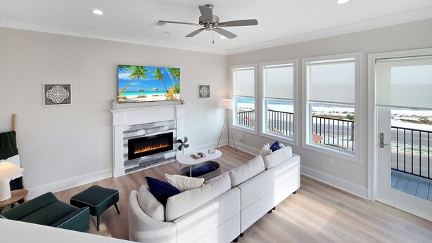 A modern living room with a fireplace, a ceiling fan, and large windows overlooking a balcony. - Seaside Villa - Gulf Breeze with an Ocean View! (Long Beach)