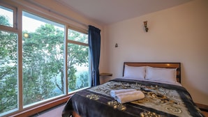A hotel room with a large window, a bed with a patterned bedspread, and a small wall decoration. - Samrakshitha Suits  (Kotagiri)