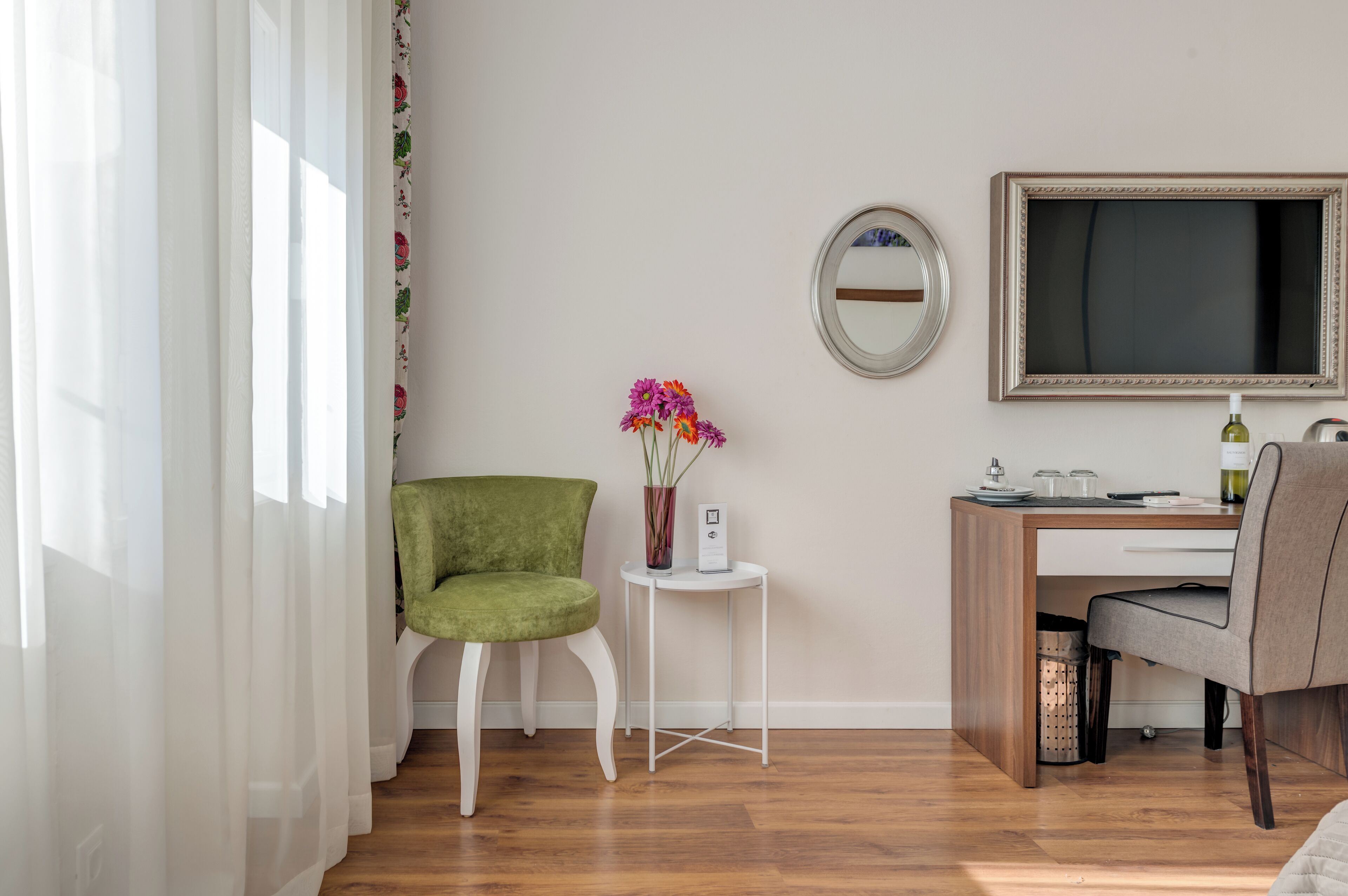 Une pièce avec un bureau en bois, une chaise verte, une chaise blanche, une petite table d’appoint sur laquelle se trouve un vase de fleurs, un miroir rond et un téléviseur fixé au mur.