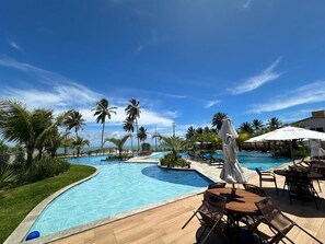 Zona de piscina en un resort, con piscina curva, sillas de descanso, sombrillas y palmeras.