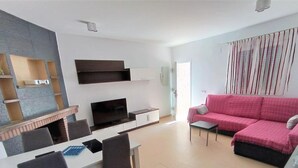 A modern living room with a flat-screen TV, a pink sofa, and a dining area with a white table and chairs.