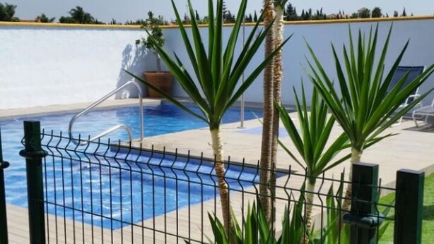 A swimming pool with a metal fence, palm trees, and a clear sky. - Casa Antonio III 113 (Conil de la Frontera)