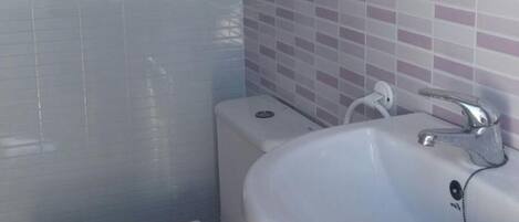 Une salle de bain avec un lavabo blanc, un miroir et des toilettes. Les murs sont recouverts de carreaux, dans une combinaison de couleurs blanche et rose.