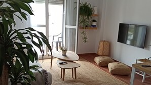 Una sala de estar con una planta grande, una mesa redonda, un sofá y un televisor montado en la pared.