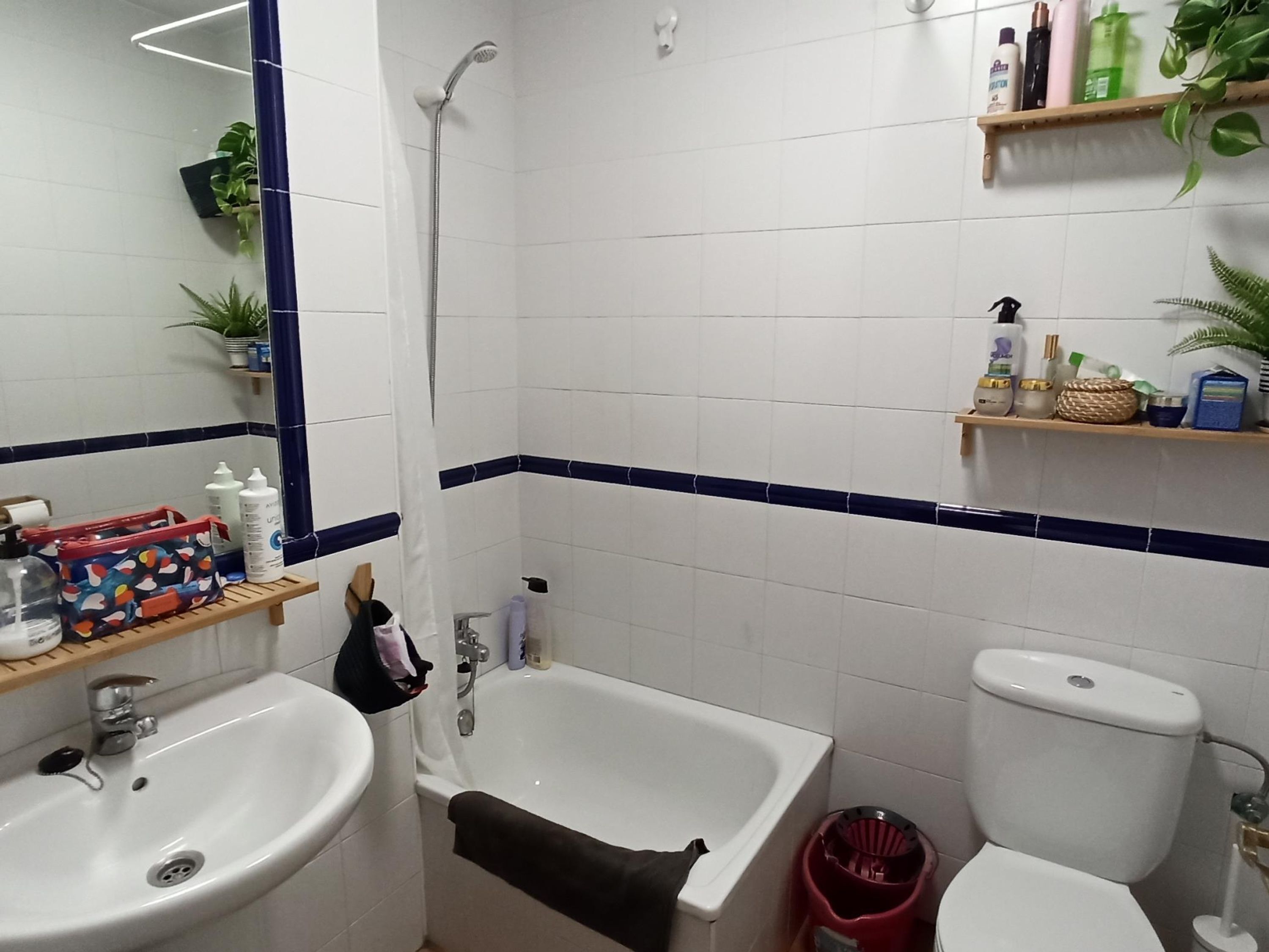 Un baño con lavamanos, bañera y retrete. Hay estantes con artículos de aseo y un espejo.