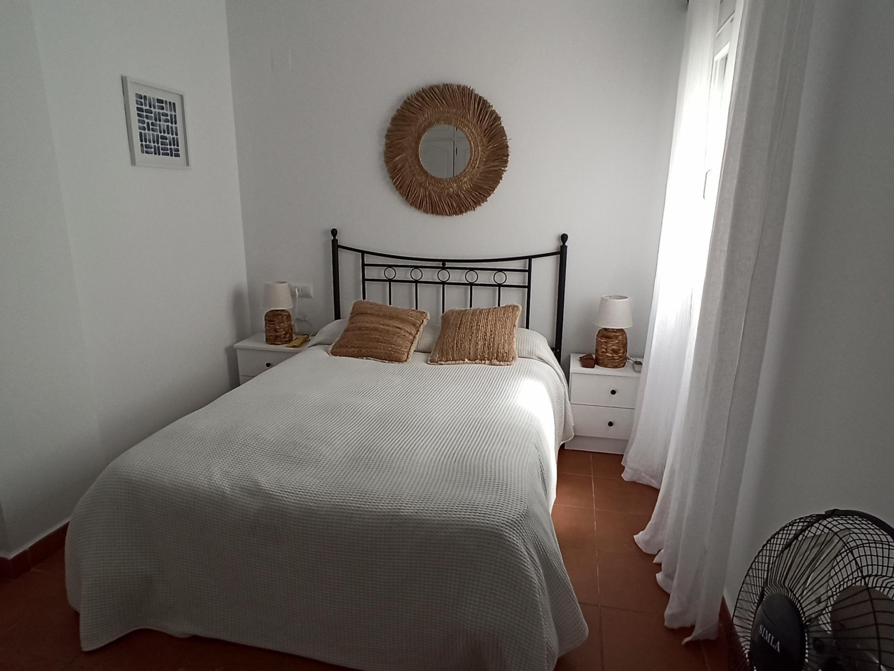 A neatly arranged bedroom with a bed, two bedside tables, a wall-mounted mirror, and a fan.