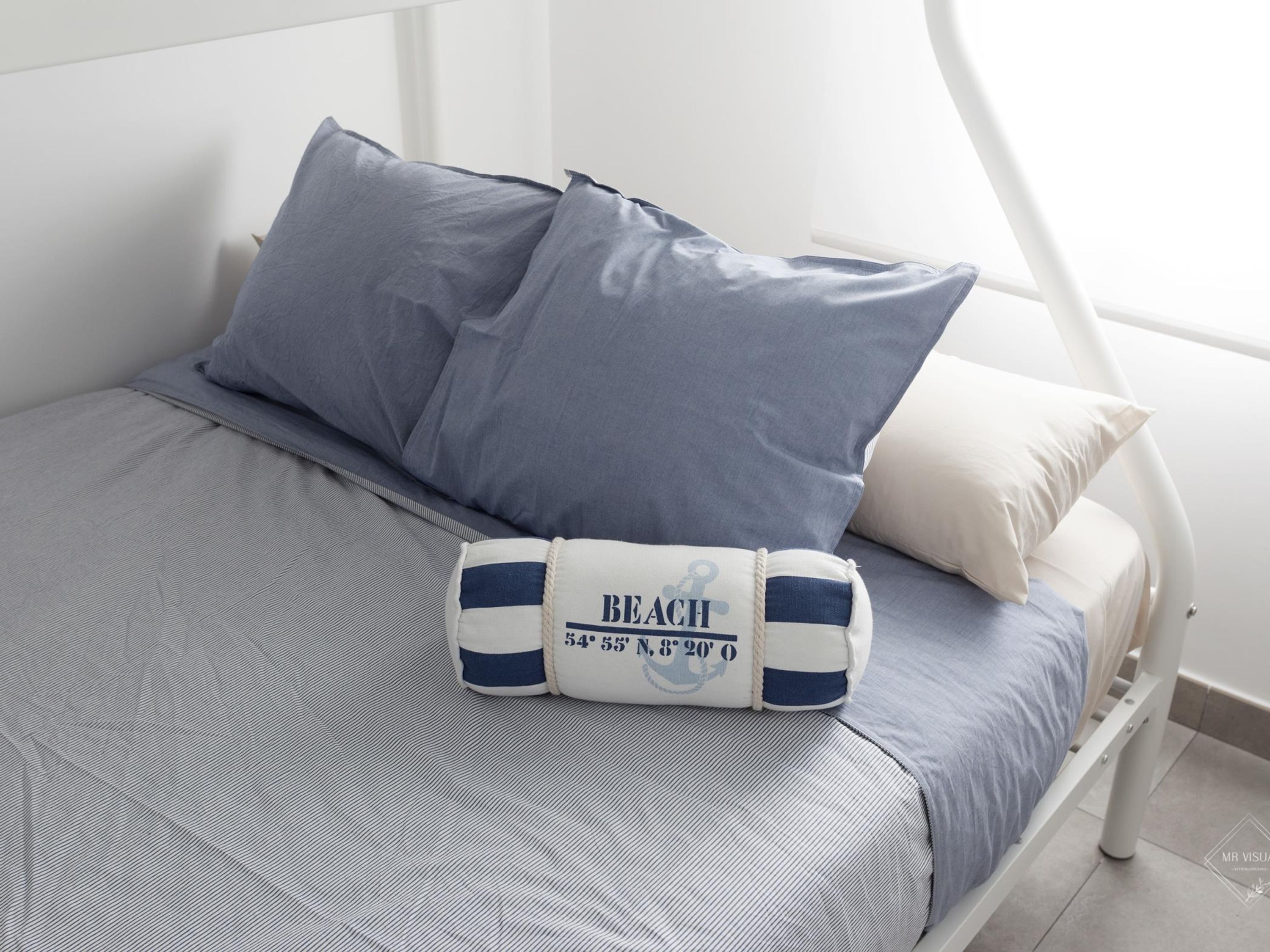 Ein Bett mit einer grau-weißen gestreiften Tagesdecke, zwei graue Kissen und ein weißes Kissen mit blauem Streifen.