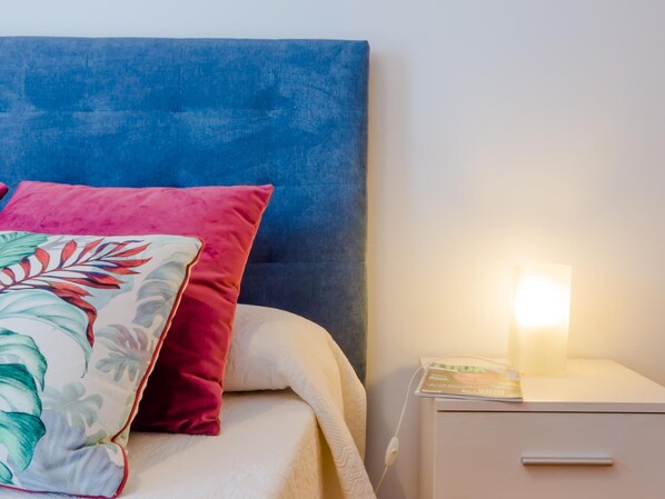 A neatly made bed with a blue headboard, a white bedside table with a lamp, and a nightstand with a book.