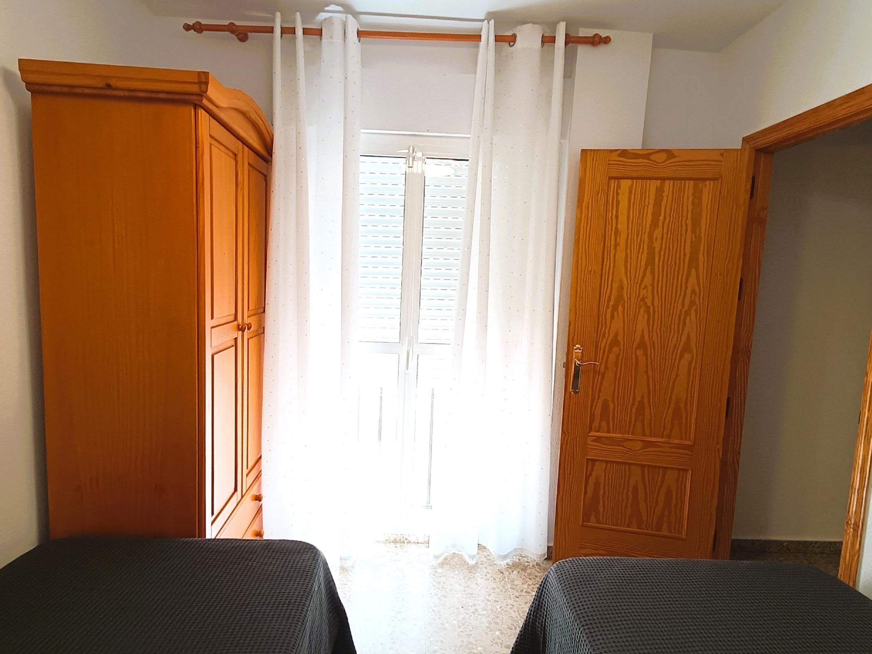 A room with two beds, a wooden wardrobe, a door, and a window with white curtains.