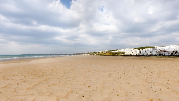 Ein weiter Sandstrand, bewölkter Himmel und weiße Gebäude im Hintergrund.
