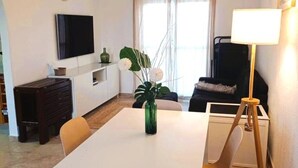 A modern living room with a white table, a flat-screen TV mounted on the wall, a dark-colored sofa, and a floor lamp. - Apartamento Canela 2536 (Conil de la Frontera)