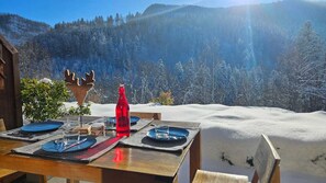 Une table dressée pour deux personnes, avec une bouteille rouge, des assiettes et des couverts, donnant sur un paysage montagneux enneigé.