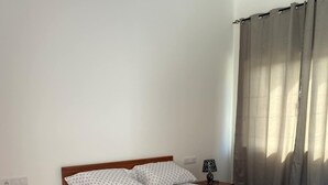 A bedroom with a wooden bed, white patterned bedding, a nightstand with a lamp, and a window with curtains. - Laranjinha Rooms (Paul)
