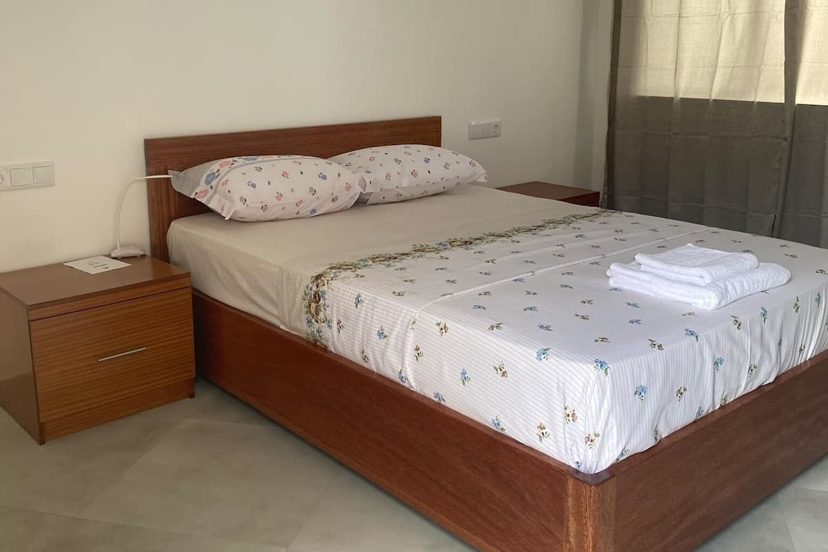 Quarto com cama de madeira, mesa de cabeceira, tomada e janela com cortinas.