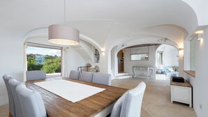 Un comedor amplio con mesa de madera y sillas blancas, techo abovedado y vista a zonas verdes.