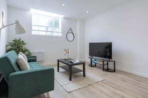 Ein modernes Wohnzimmer mit einer Couch, einem Couchtisch, einem Fernseher auf einem Schrank, einer Stehlampe und einem großen Fenster.
