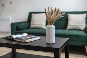 A room with a green sofa, a white vase with dried plants, and a magazine on a black table.