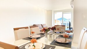 Un comedor con una mesa dispuesta para cuatro personas, rodeada de sillas. Se observa un sofá al fondo y una ventana con vistas a edificios.