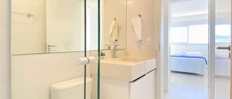 A modern bathroom with a glass shower enclosure, a toilet, a sink with a mirror, and a bed visible through an open door.