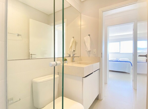 Un baño moderno con cabina de ducha de vidrio, inodoro, lavabo con espejo y una cama visible a través de una puerta abierta.