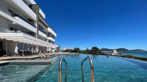 Una piscina con barandillas de metal, una cubierta de madera y vistas a un edificio con balcones y un cielo despejado.