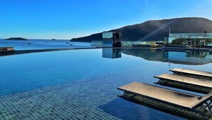 Ein Infinity-Pool mit klarem Wasser, das Himmel und umliegende Landschaft spiegelt, ein modernes Gebäude mit Glasfassaden und Blick auf das Meer mit Booten.
