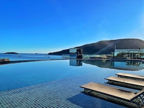 An infinity pool with clear water reflecting the sky and surrounding landscape, a modern building with glass walls, and a view of the sea with boats. - Espelho das Águas Residencial (Florianópolis)