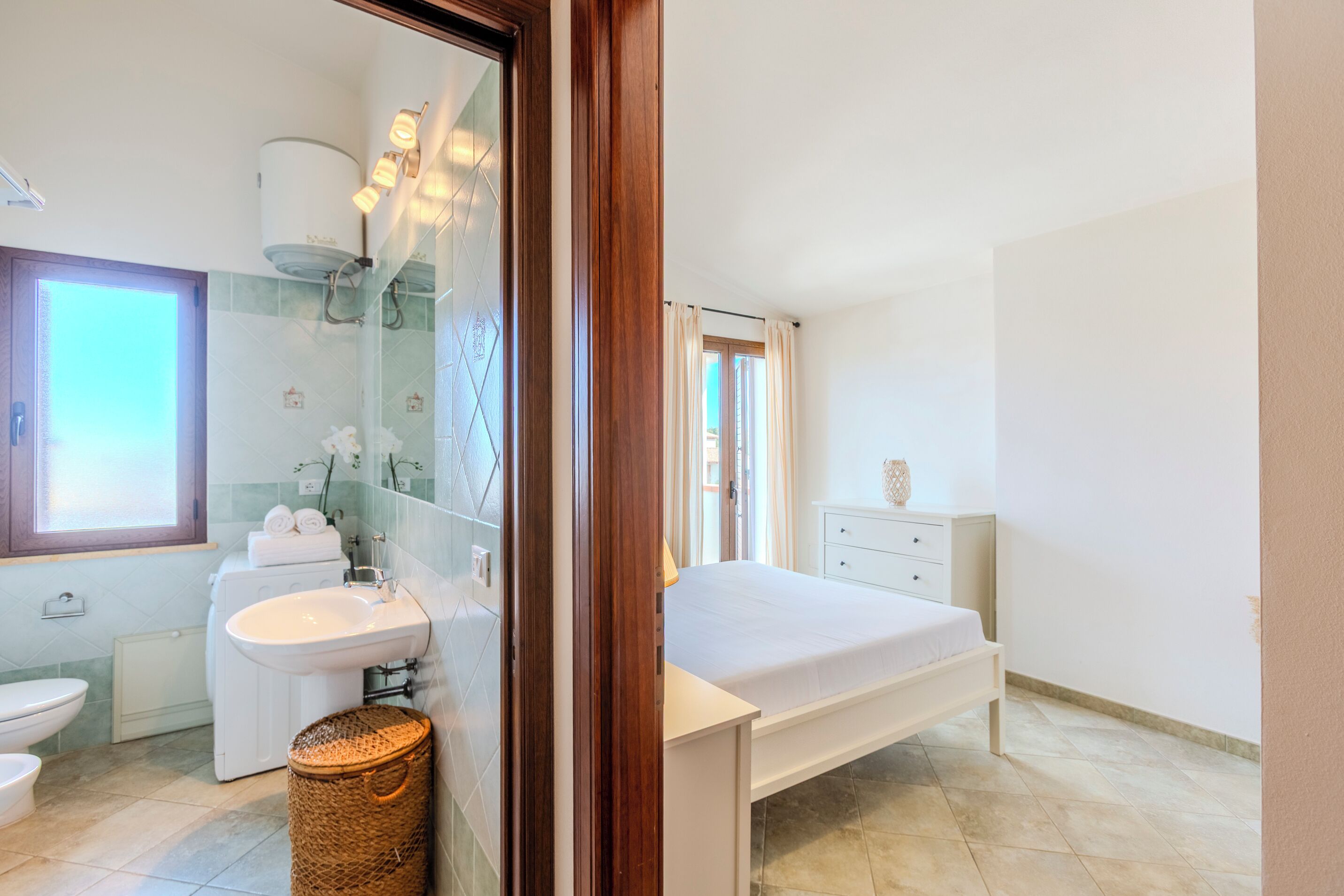 A modern bathroom with a toilet, sink, and shower. There is a bed with a white mattress and a bedside table with a lamp.