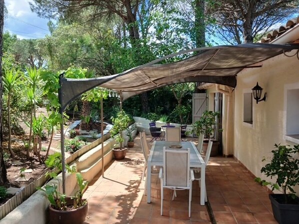 Un patio con mesa y sillas, rodeado de plantas en macetas y una zona con sombra.