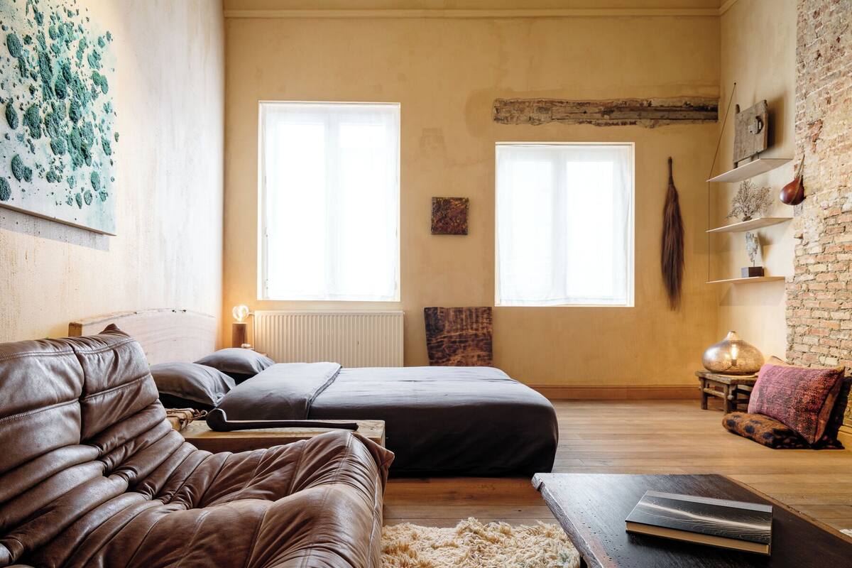 A cozy bedroom with a large bed, a leather sofa, and a wooden coffee table. There are two windows providing natural light, and a brick wall adds character to the room.