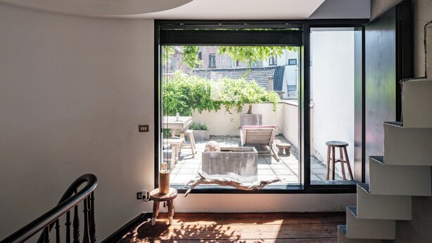 A modern interior with a staircase leading to a balcony, a wooden floor, and a view of a garden and buildings outside. - Verne Dreams (Ghent)
