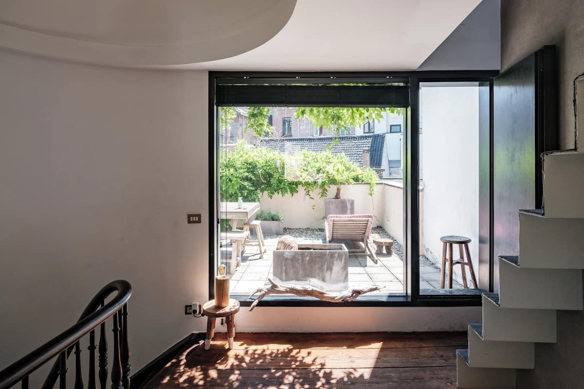A modern interior with a staircase leading to a balcony, a wooden floor, and a view of a garden and buildings outside.