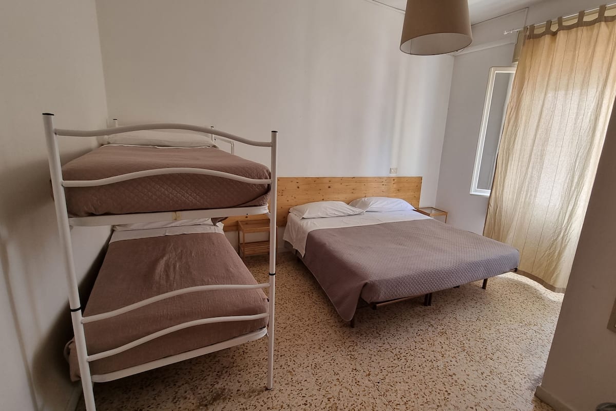 Ein Zimmer mit Etagenbetten, einem Einzelbett, einem hölzernen Kopfteil, einem kleinen Nachttisch und einem Fenster mit Vorhängen.