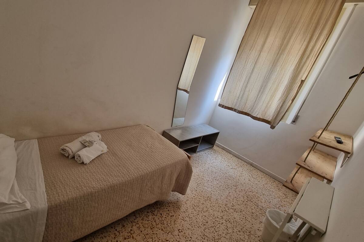 Ein kleines, ordentlich eingerichtetes Schlafzimmer mit Bett, Nachttisch, Spiegel und einem Fenster mit Vorhängen.