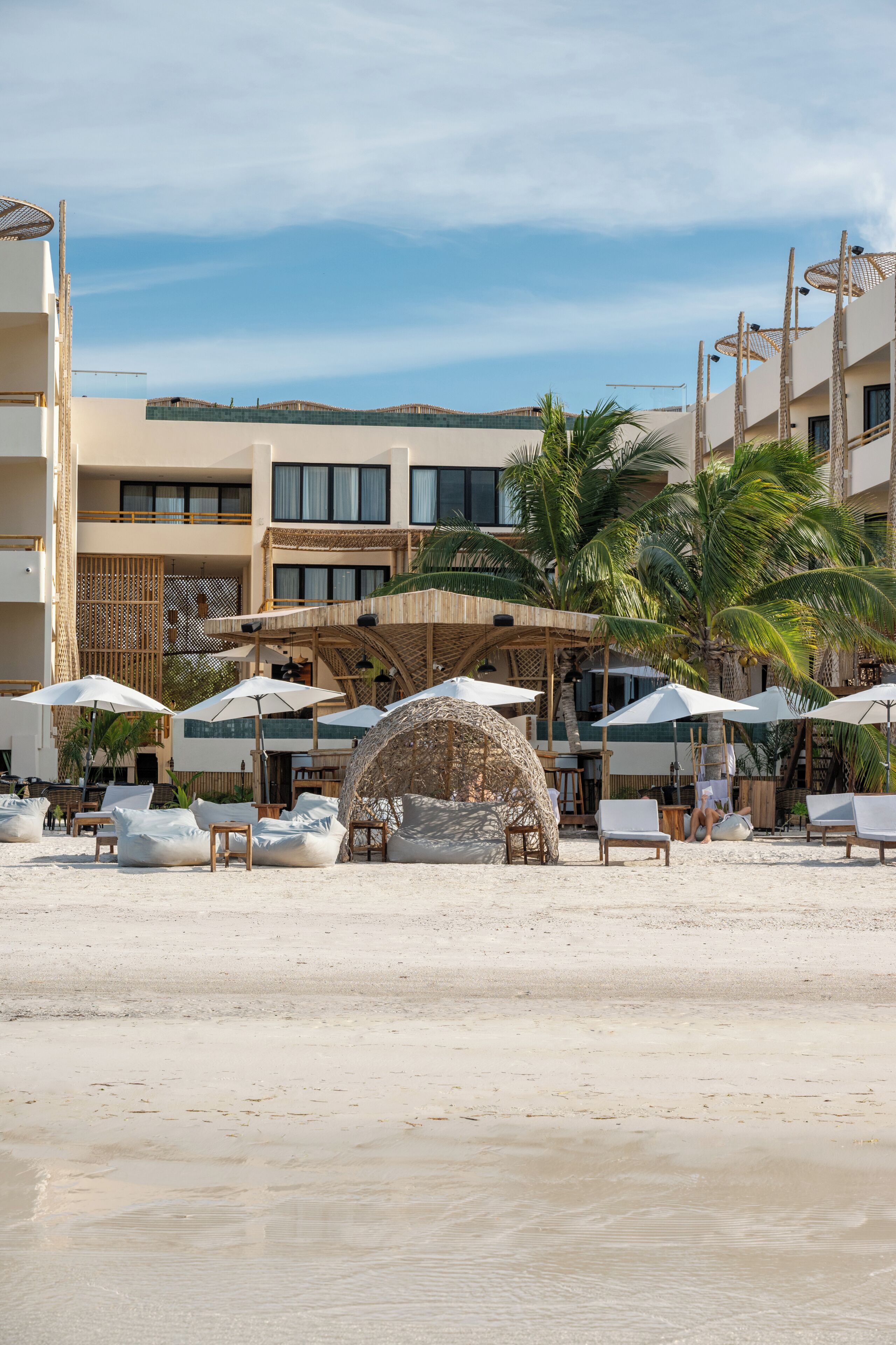 Un hotel frente a la playa con mesas al aire libre, sombrillas y palmeras.