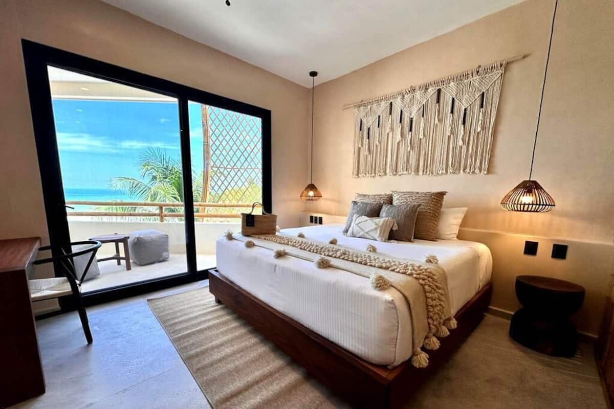 Une chambre avec un grand lit, une décoration murale en macramé et une vue sur l’océan par une porte coulissante.