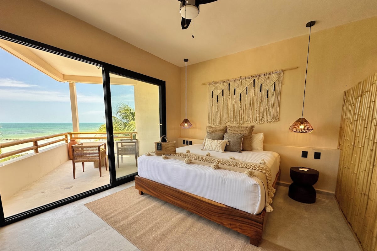 Une chambre avec un grand lit, une vue sur l’océan, un balcon et une décoration murale en macramé.