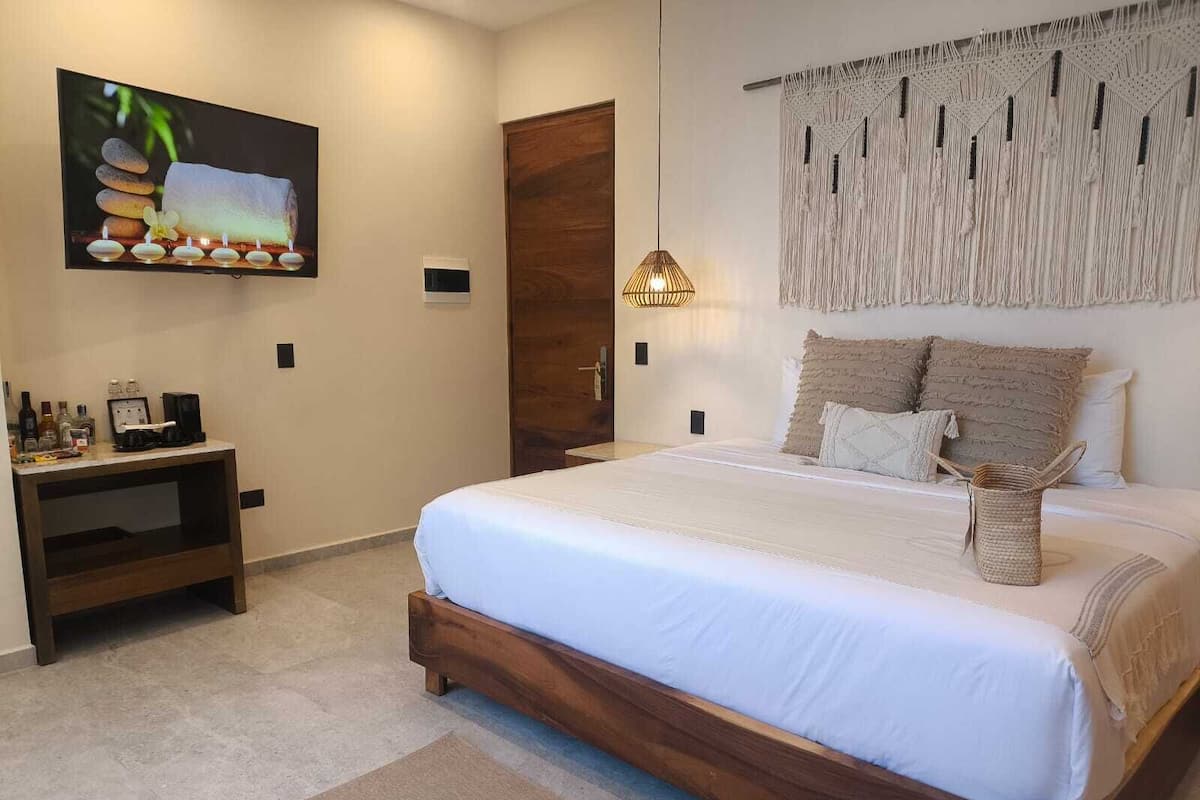 Une chambre avec un grand lit, une table de chevet en bois avec une lampe, un téléviseur fixé au mur et une décoration murale en macramé.