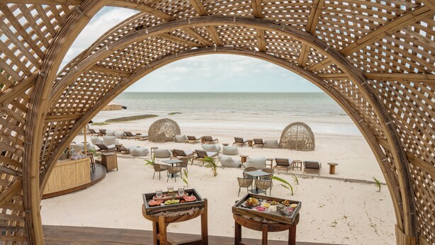 A beachside lounge with a wooden arch structure, lounge chairs, and tables with food trays. - Ana y Jose Holbox Boutique All Inclusive Hotel (Isla Holbox)