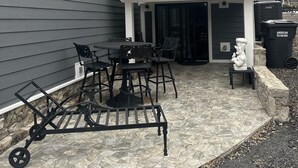Et toetagers hus med balkon, udendørs siddeområde med stole og bord samt en stenbelagt terrasse.
