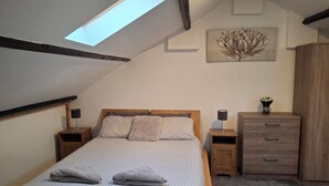 Une chambre avec un plafond en pente, un puits de lumière, un lit avec une literie à rayures, deux tables de chevet avec des lampes, une commode en bois et une œuvre d’art accrochée au mur.