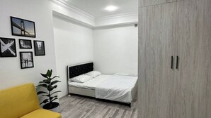 Een slaapkamer met een bed, een gele slaapbank, een houten bijzettafeltje, een kledingkast en een potplant.