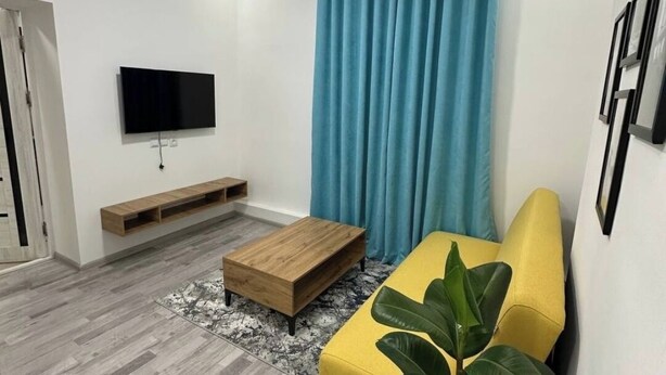 Een moderne woonkamer met een flatscreen televisie, een houten salontafel, een gele bank en een grote plant.