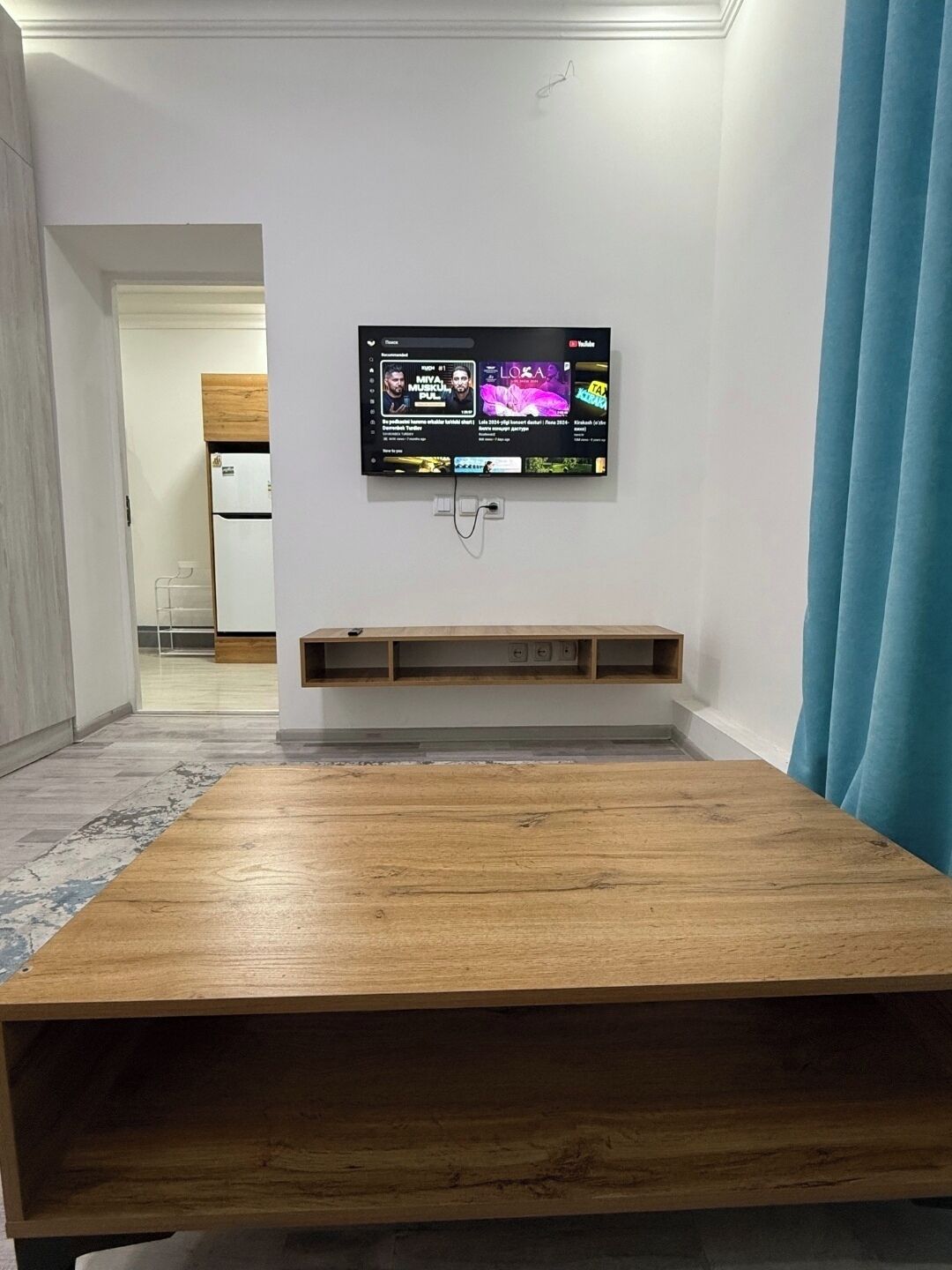 Quarto com uma mesa de madeira, TV fixada exibindo videoclipes e uma área de cozinha ao fundo.