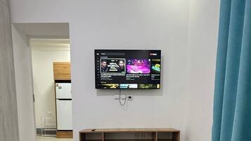 Quarto com uma mesa de madeira, TV fixada exibindo videoclipes e uma área de cozinha ao fundo.