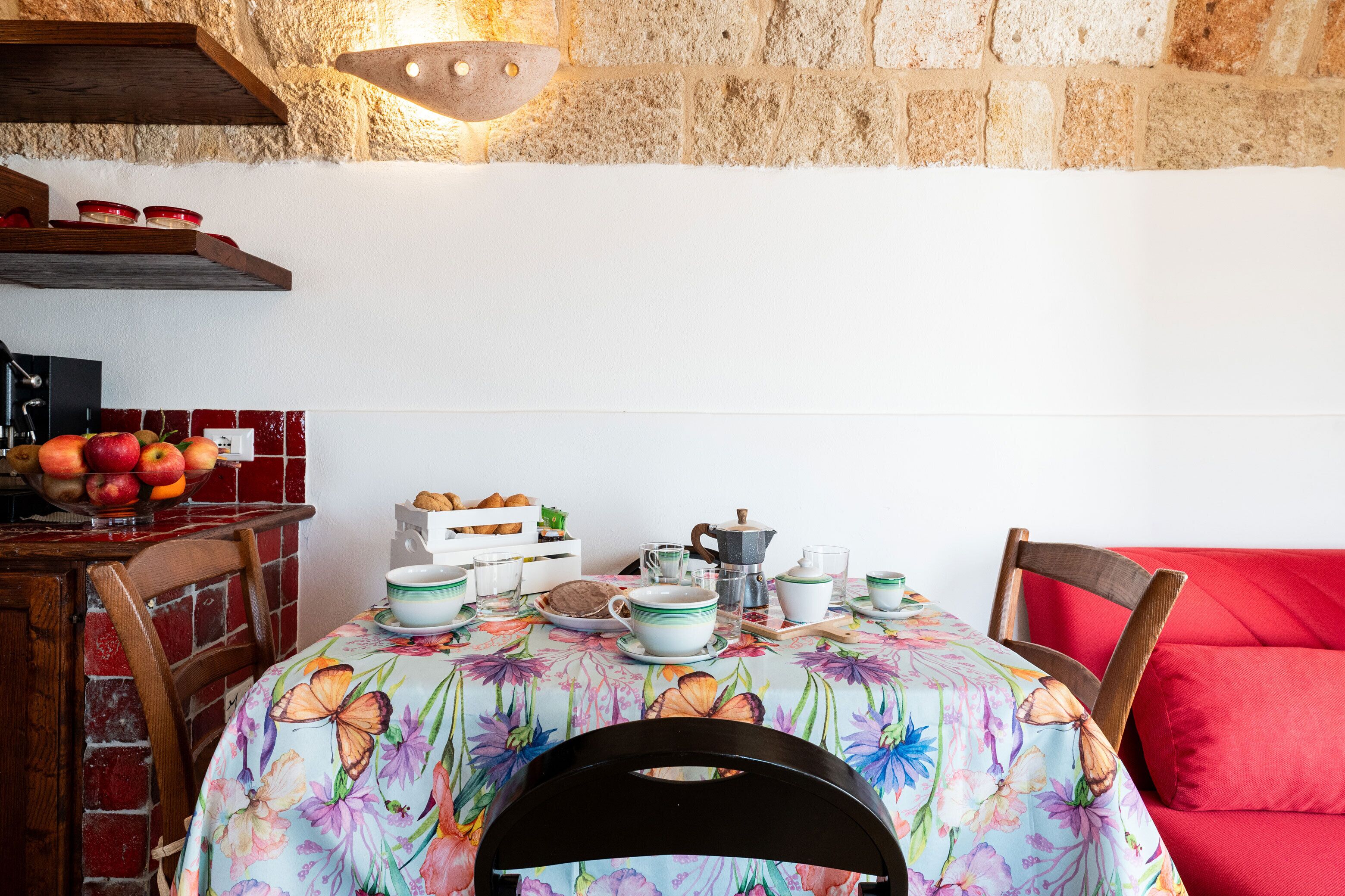 Un comedor con una mesa preparada para dos personas, con mantel floral, sillas de madera y un sofá rojo.