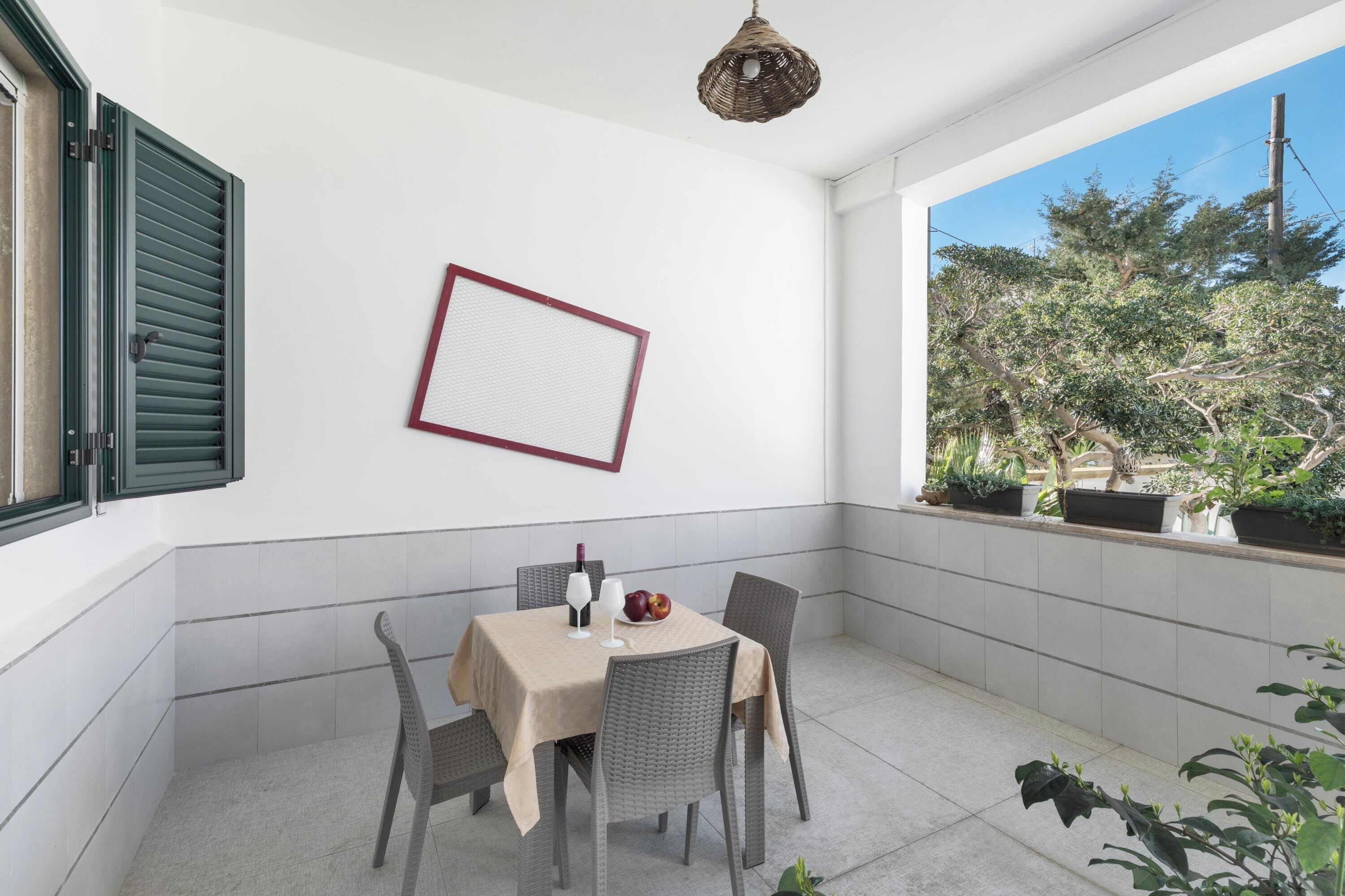 A dining area with a table set for two, a wicker chair, a hanging light fixture, and a view of trees through a window.